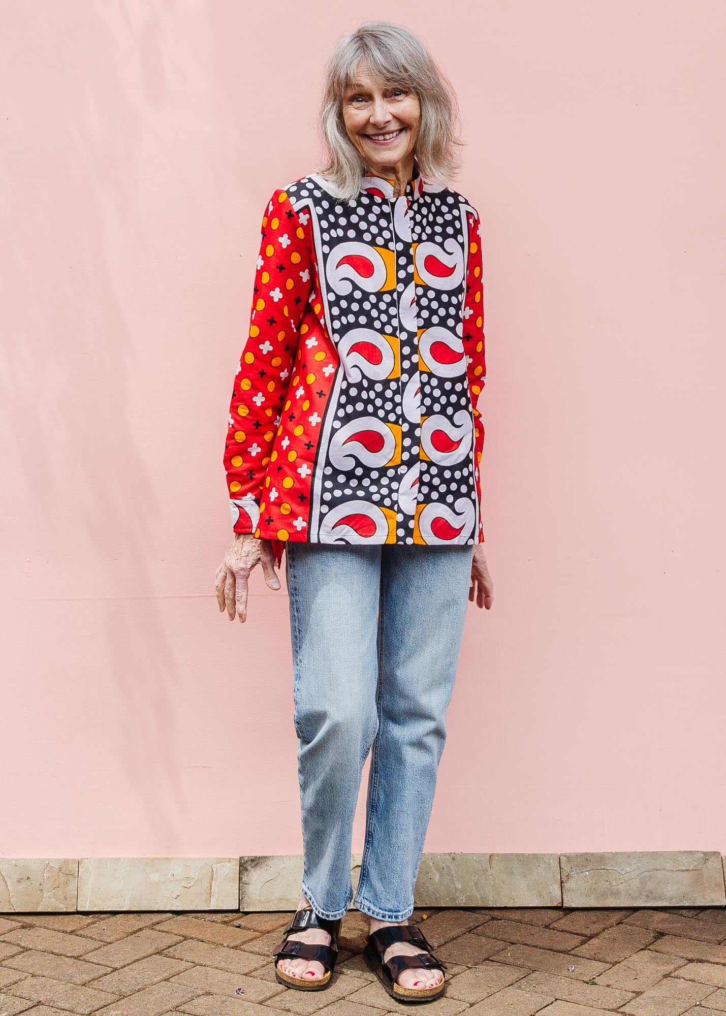 The model is wearing red, white, black and orange mixed pattern long sleeved kanga shirt